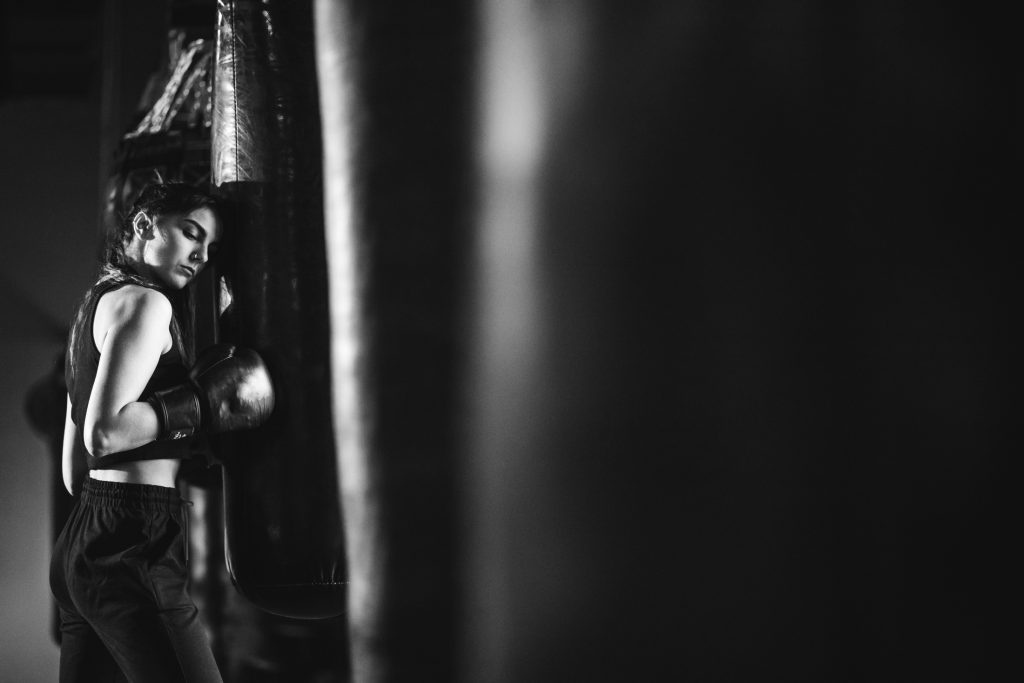 Black and white photo of a woman at the gym leaning against a punching bag.